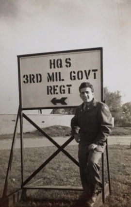 Sgt. Toriello in front of sign that says 3rd MIL GOVT REGT with an arrow pointing left