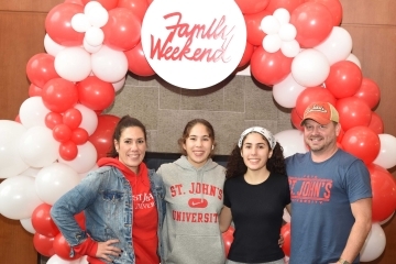Two daughters standing with parents infront of SJU Family Weekend balloon arch