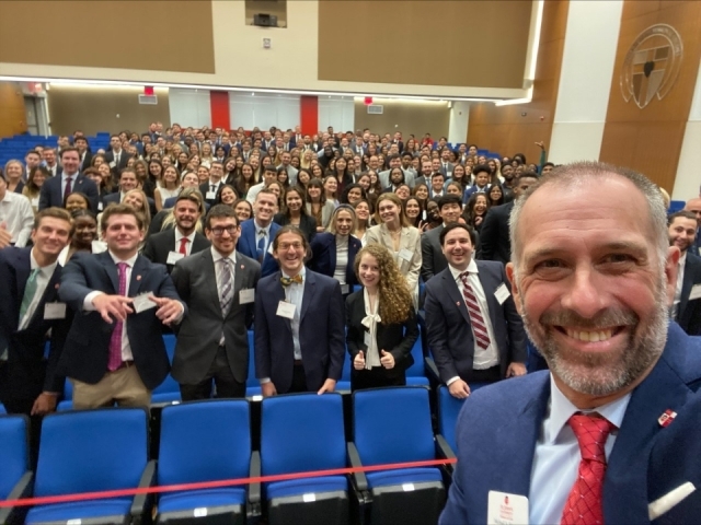 Dean Michael A. Simons stands in front of the room and takes a selfie with students standing and smiling in the background.