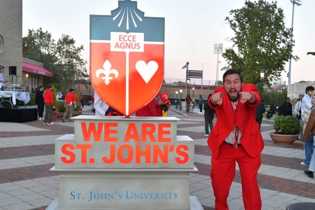 Make in red suit pointing at the camera while standing next to the St. John's University statue 