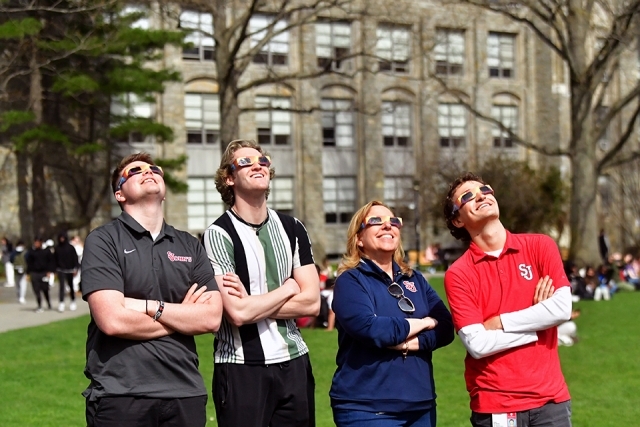 Four individuals wearing solar eclipse glasses looking up at the sky