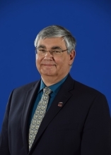 Professional head shot of Joseph Brocavich, standing in front of a blue background, wearing a blue blazer, teal collared shirt, and multi-colored tie. He wears glasses, has salt and pepper hair, and is smiling.