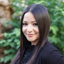 Portrait of a woman standing outdoors with a soft-focused greenery background. She has long, straight dark hair, is smiling gently, and is wearing a black top with lace detailing at the collar. 