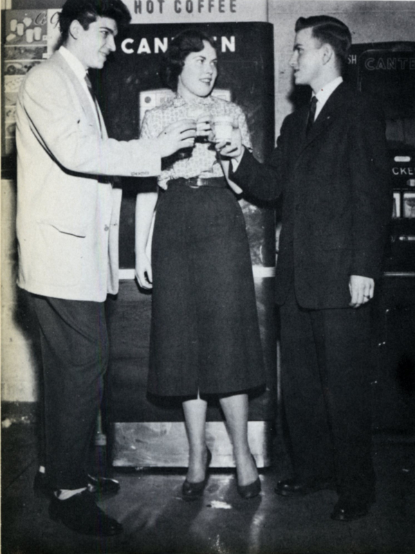 Three students drinking coffee in front of the cafeteria vending machines.