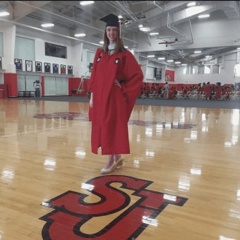 A woman in a red graduation gown and cap stands proudly on the St. John’s University gym floor, smiling beside the large “SJ” logo as other graduates gather in the background.