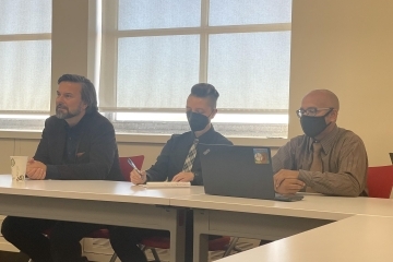 Three faculty members sitting at a table listening to others speak in room