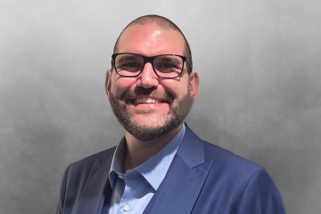 St. John's Law alumnus Ryan Bravata, wearing a blue blazer and blue dress shirt with an open collar, smiles facing forward.