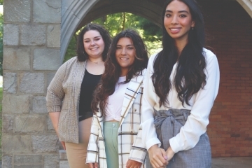 Three Rose DiMartino student fellows stand by a stone arched building entrance.
