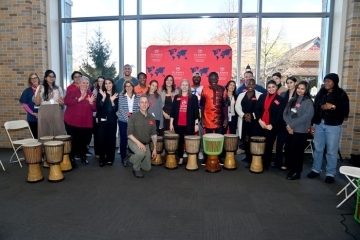 A large group of students, staff, and guest drummers pose together in front of the St. John’s University International Education backdrop with hand drums arranged in front of them.