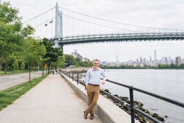 Hunter Igoe stands on a tree -lined riverside promenade in new York City with a bridge and the city's skyline in the background.