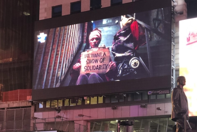 Screen displaying image of a young woman holding a sign that says "We Want a Show of Solidarity"