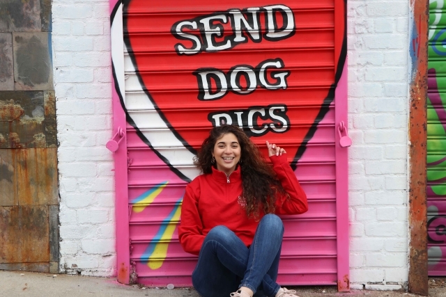 Mural with Send Dog Pics and Ariana sitting in front of it 