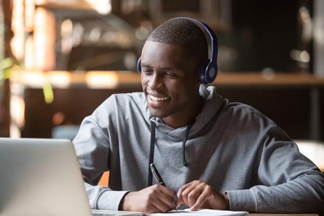 Young man sitting at laptop with headphones and writing on a paper