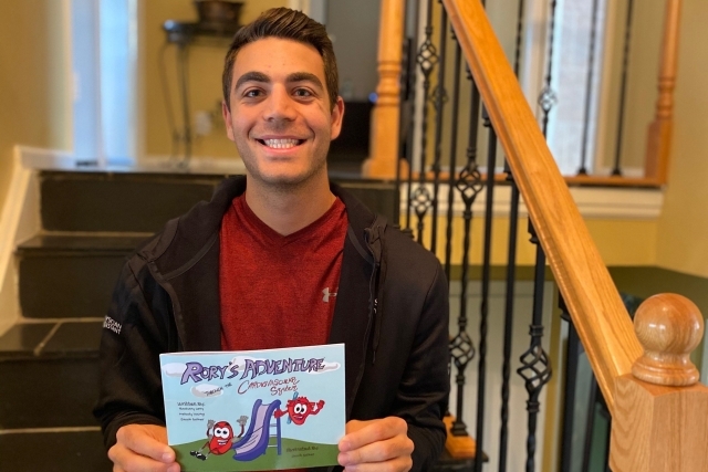 Zachary Levy sitting on a set of stairs holding his book "Rory's Adventures of the Cardiovascular System"