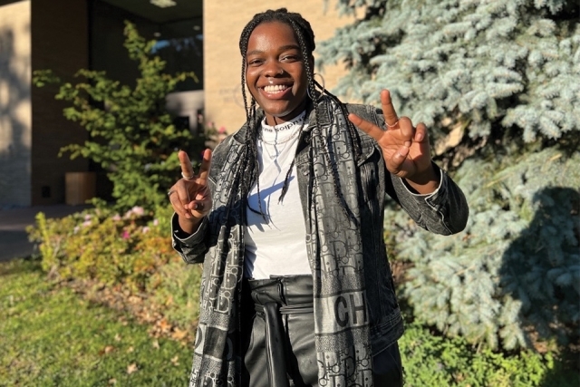 St. John's Law student camille castle wearing a dior outfit with a white shirt, dark jacket, dark pants, and a scarf smiles with both her hands raised and her fingers making peace signs