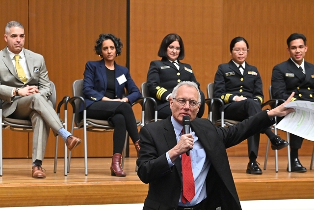Moderator speaking into a microphone with panelists behind him onstage