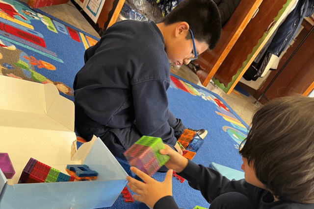 Two children sit on a colorful classroom rug, building with translucent magnetic tiles. One child reaches into a box of tiles while the other arranges pieces on the floor.