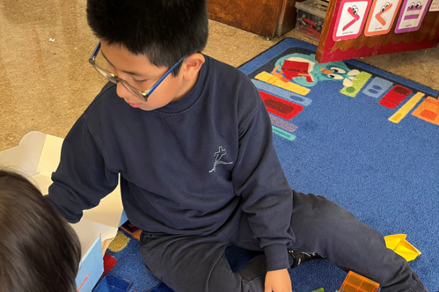 A child wearing glasses sits on a colorful classroom rug, reaching toward a box of magnetic tiles while playing with another student.