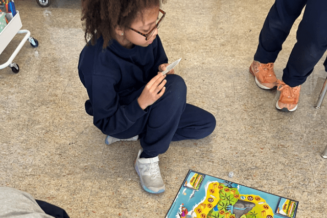 A child sits on the classroom floor reading a card while playing a board game. The board and game pieces are spread out on the tile floor.