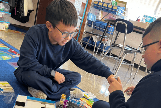 Two children sit on the floor of a classroom, working together with small building pieces. One child holds a piece while the other examines the set.