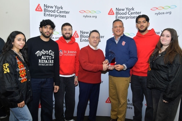 “Group of seven people standing in front of a New York Blood Center Enterprises backdrop, with two individuals in the center holding the Milestone Award.”