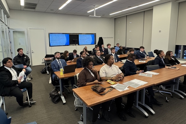 Wide view of a modern classroom with students in professional attire attending a finance or business lecture.
