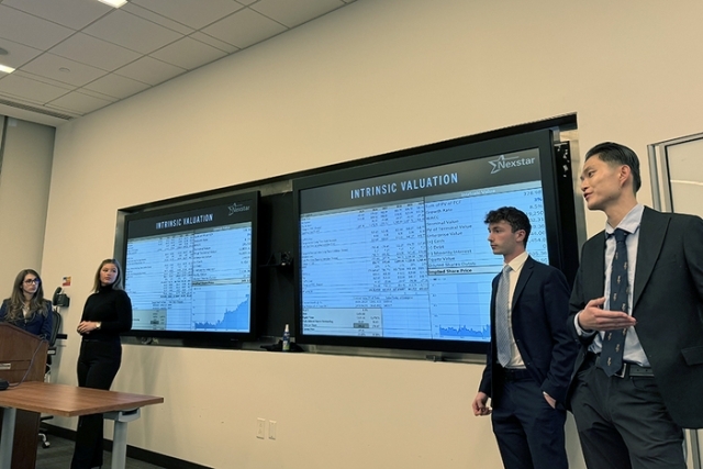 Students presenting financial valuation data displayed on multiple screens during a classroom presentation.
