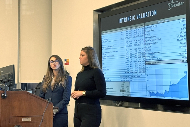 Students in professional attire presenting a healthcare sector analysis in a classroom using a large digital screen.