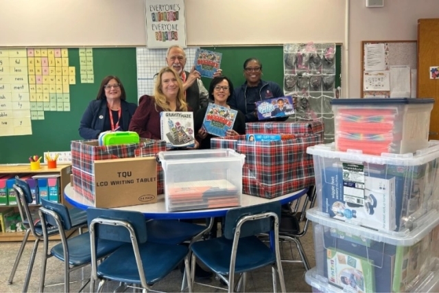 Five adults smile while standing behind a classroom table filled with large plastic bins, boxes of educational toys, and supplies. A chalkboard and classroom materials are visible in the background.