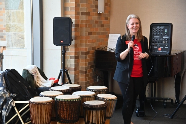 A speaker holding a microphone stands beside a row of hand drums and sound equipment at a campus cultural celebration.