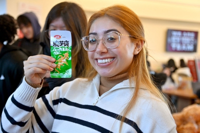 A smiling student holds up a packaged snack from an international table during a campus cultural event.