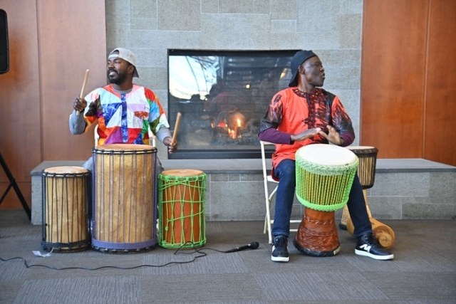 Two musicians perform on traditional drums in front of a fireplace at a campus cultural program.