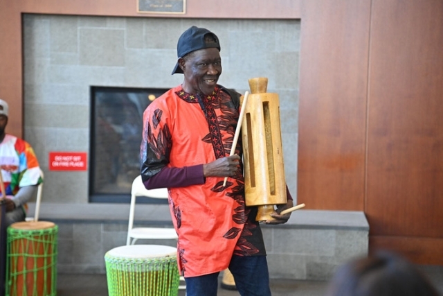 A guest performer holds a wooden slit drum and smiles while demonstrating it during a cultural drumming session.