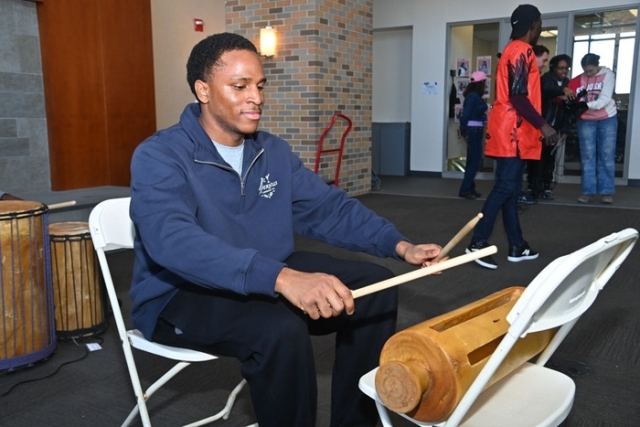 A student sits and plays a wooden slit drum with mallets during a drumming activity at a university event.