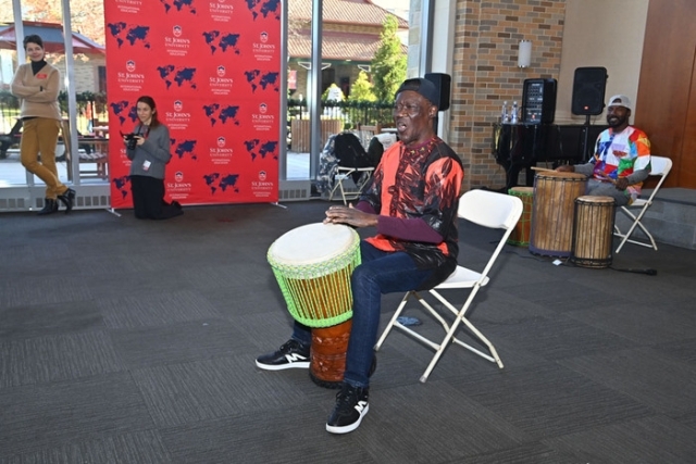 A guest performer leads a drumming demonstration while seated, with another drummer playing in the background during a campus event.