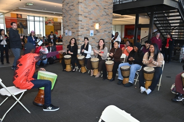 Participants sit in a semicircle playing hand drums while an instructor at the front guides the group during a campus drumming workshop.