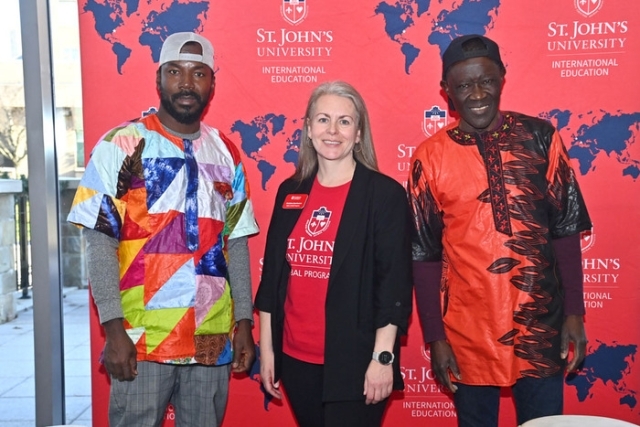 A large group of students, staff, and guest drummers pose together in front of the St. John’s University International Education backdrop with hand drums arranged in front of them.