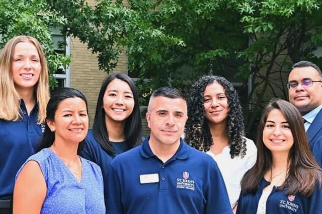 “Group of seven St. John’s University staff members smiling outdoors in blue polo shirts.”