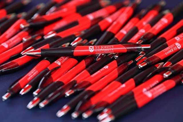 “Close-up of a pile of red and black St. John’s University branded pens on a table.”