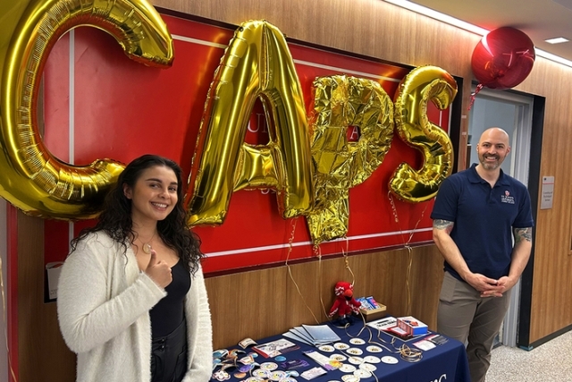 “Two St. John’s University staff members standing by a display table, with large gold balloon letters spelling ‘CAPS’ behind them.”