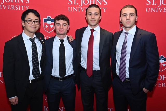 Four students or young professionals in suits posing in front of a St. John’s University step-and-repeat backdrop.