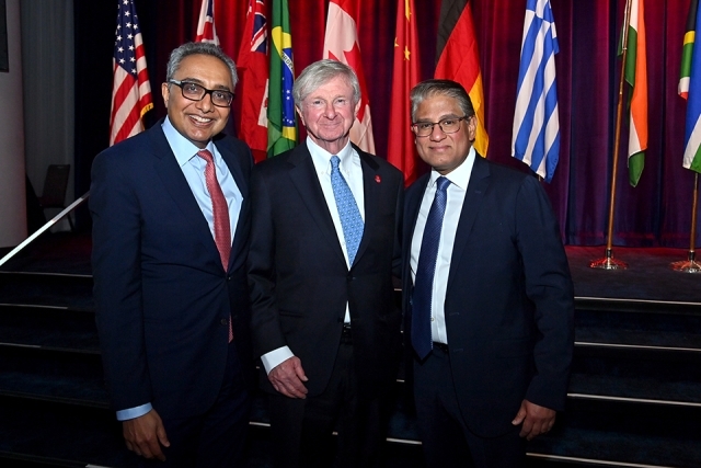 Three attendees posing together on stage with international flags displayed in the background.