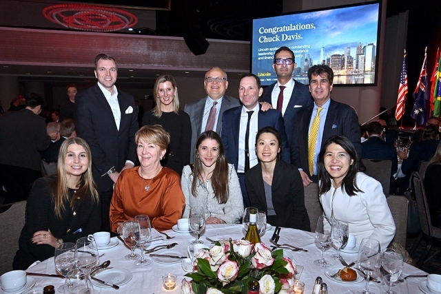 Large group of attendees posing together at a table with a presentation screen visible in the background.