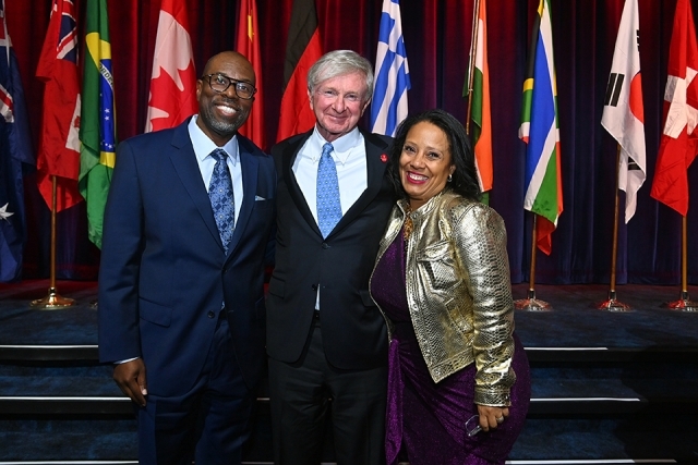 Three attendees posing together on stage with international flags displayed in the background.