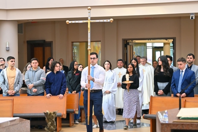 Students and clergy process down the center aisle of a chapel during a Catholic Scholars commissioning ceremony, with a student carrying a large cross at the front.