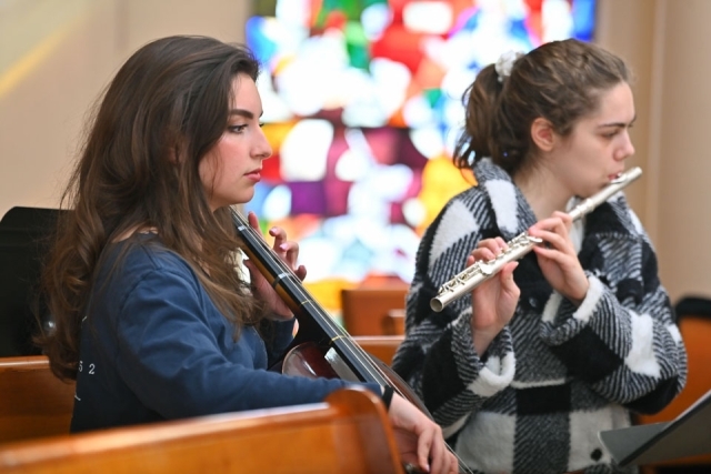 Student musicians perform during a Catholic Scholars commissioning Mass, with a violinist and flutist seated in a chapel and stained glass visible behind them.