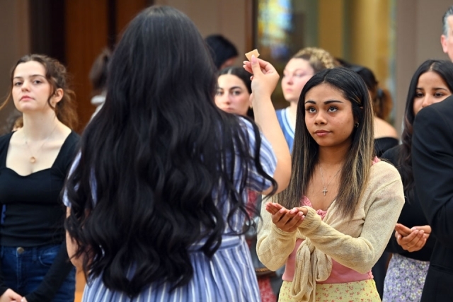 A student receives communion during a Catholic Scholars commissioning ceremony, surrounded by fellow students standing in the chapel.