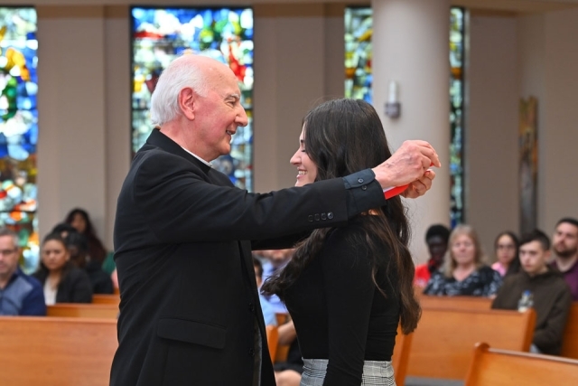 A priest places a medal over a student’s neck during a Catholic Scholars commissioning ceremony inside a chapel.