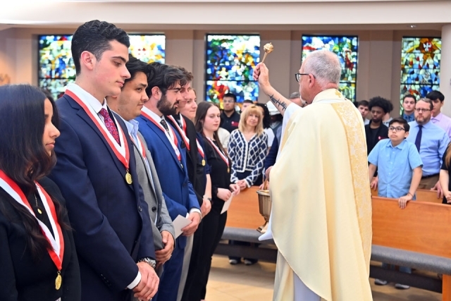 Catholic Scholars stand in a line wearing medals as a priest blesses them during a commissioning Mass in a campus chapel.