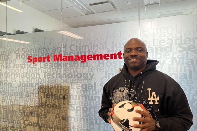 Man holding a soccer ball behind red words on a glass wall, saying "Sports Management" 